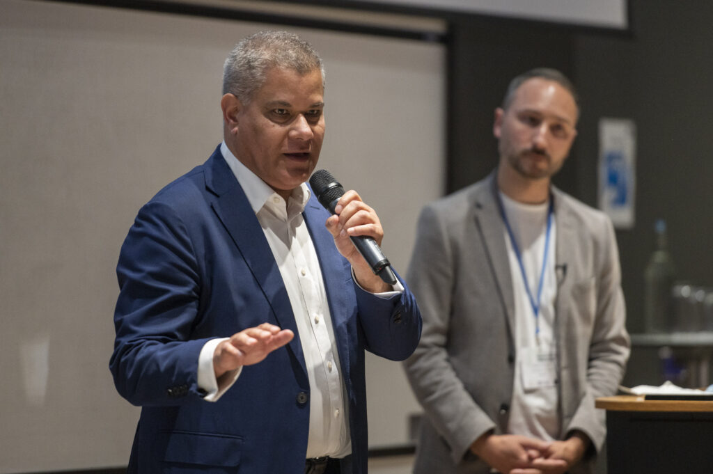 A man speaks into a microphone while another man listens in the background.