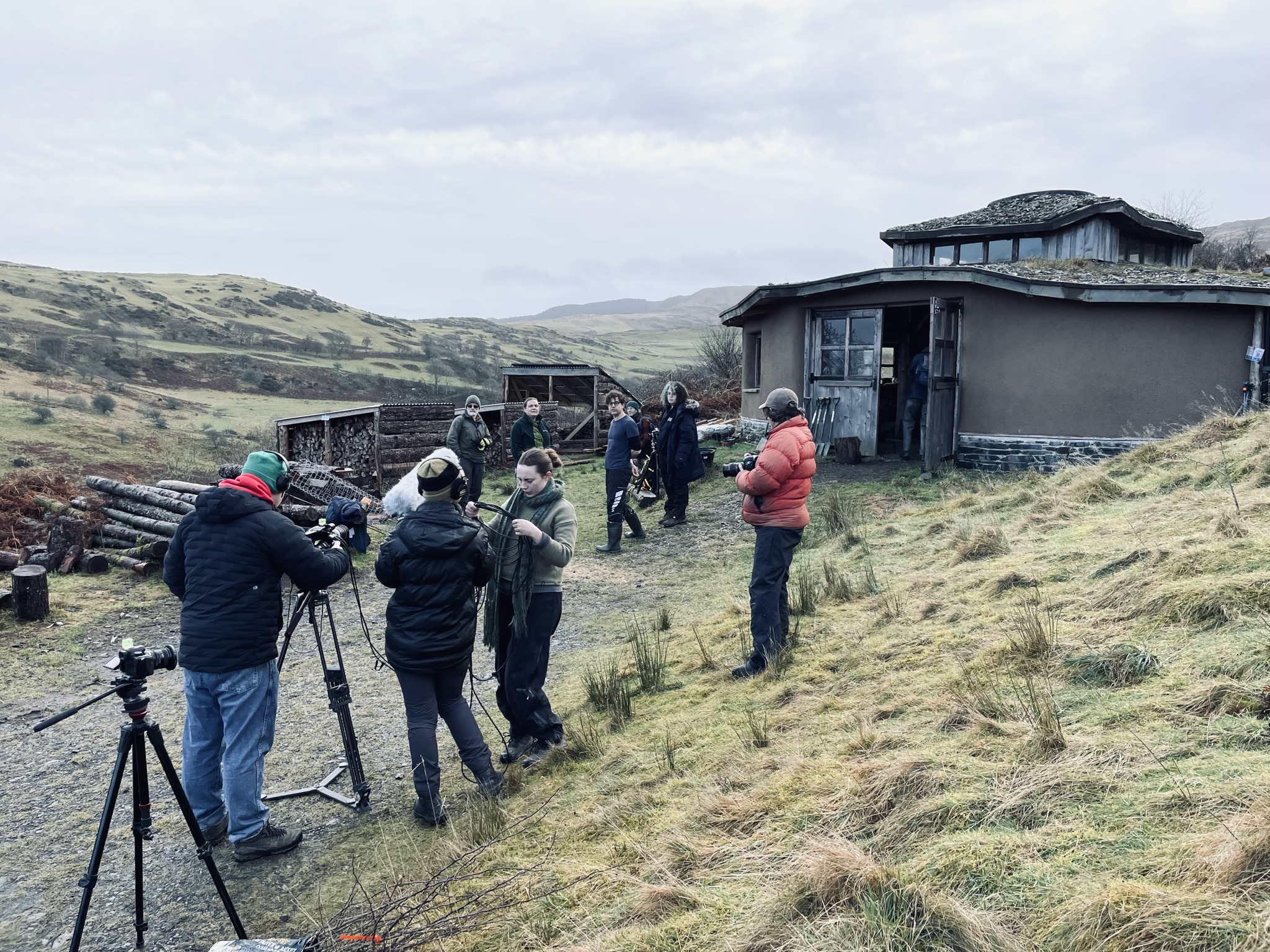 People stand in front of a house with a road leading up to it and hills on the background. They are setting up filming equipment and wearing coats and hats. There is another hill to the side of the house. 