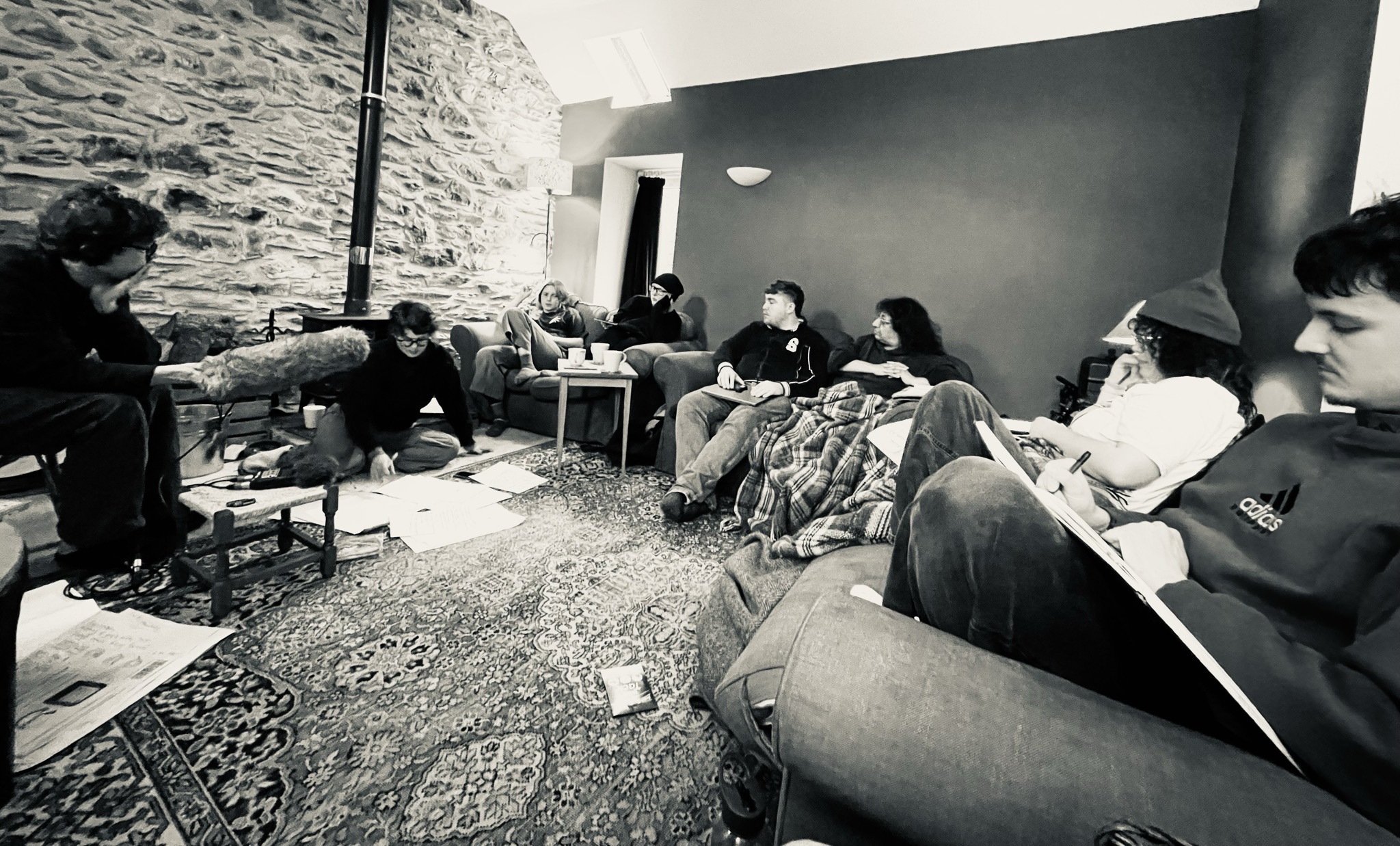 People sit on couches, chairs and on the floor as they study and have a discussion. One person is holding filming equipment. Another person is looking at papers on the floor. A person on the right side of the frame is holding a board in their lap and writing on it. 