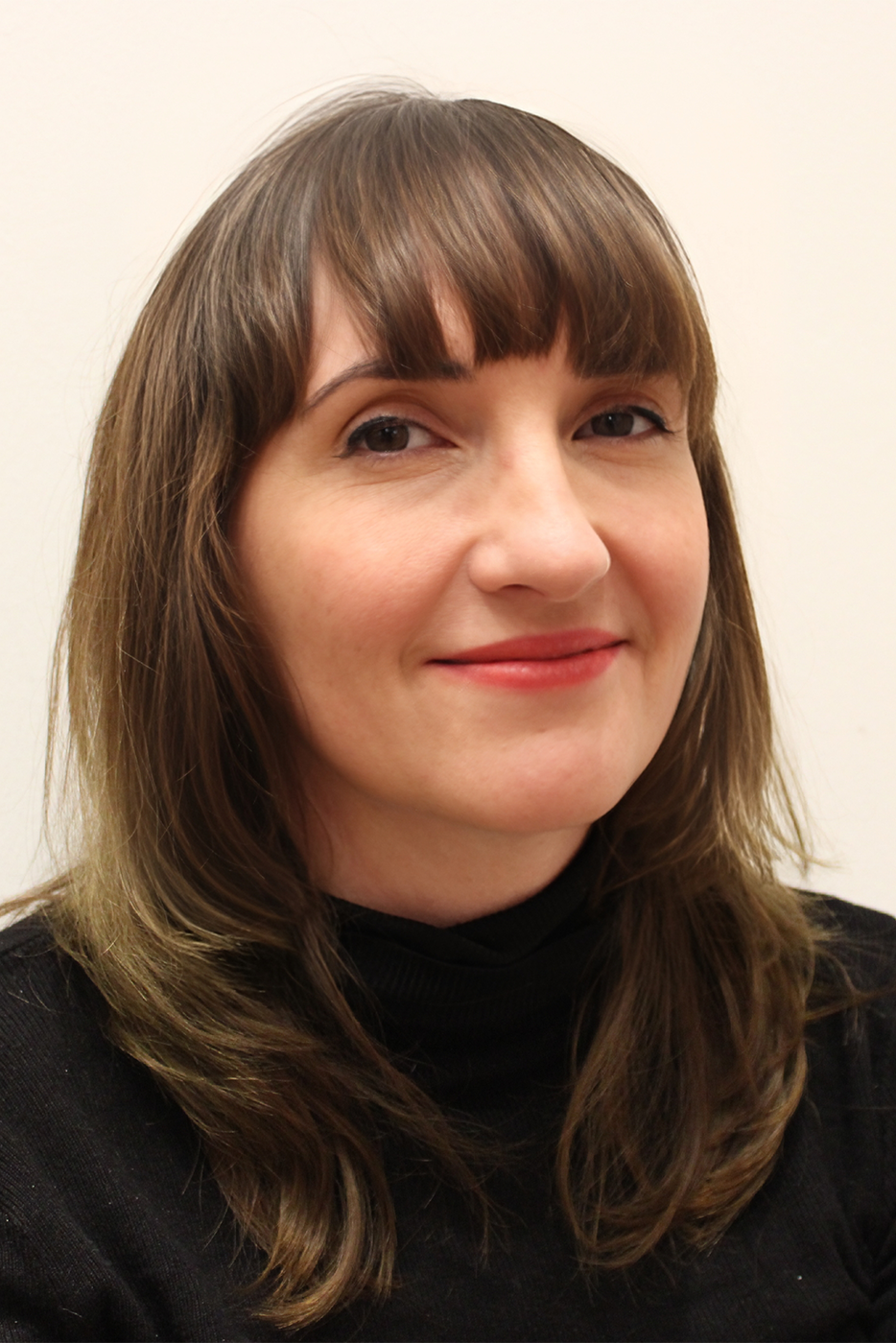 A woman smiles at the camera. She has shoulder-length hair and a fringe. She is wearing a roll-neck top. 