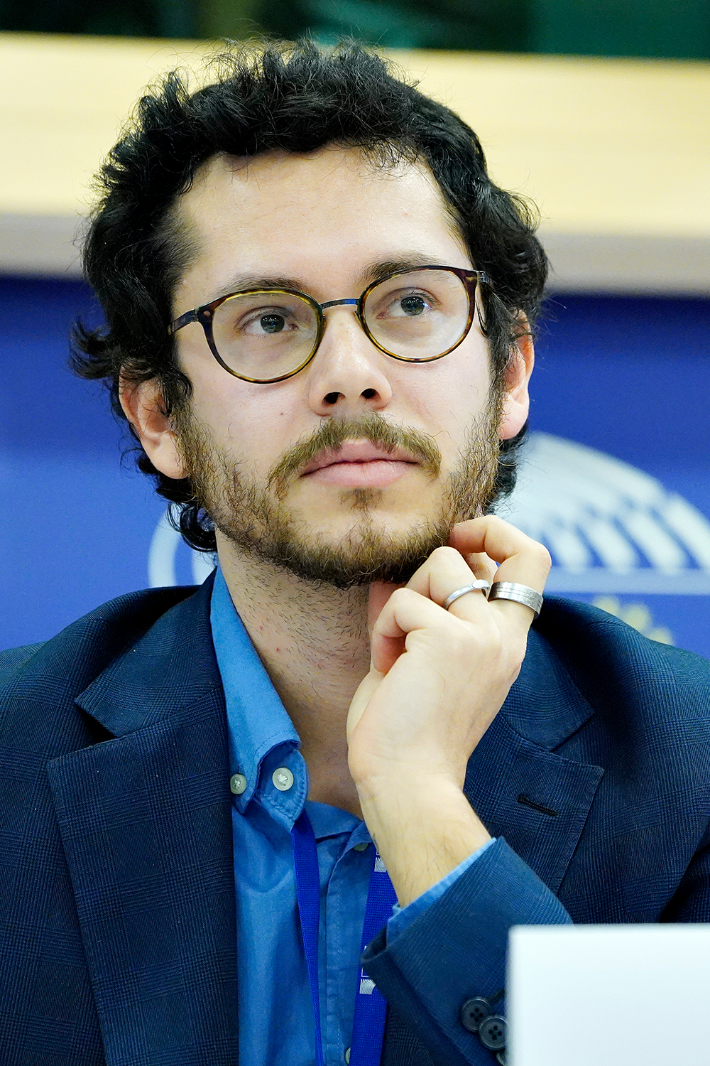 A man looks into the distance. He has short hair and a beard. He is wearing glasses, a collared shirt and a blazer. His chin is resting on one hand. He's wearing rings on two fingers. There is a backdrop with a logo behind him. 