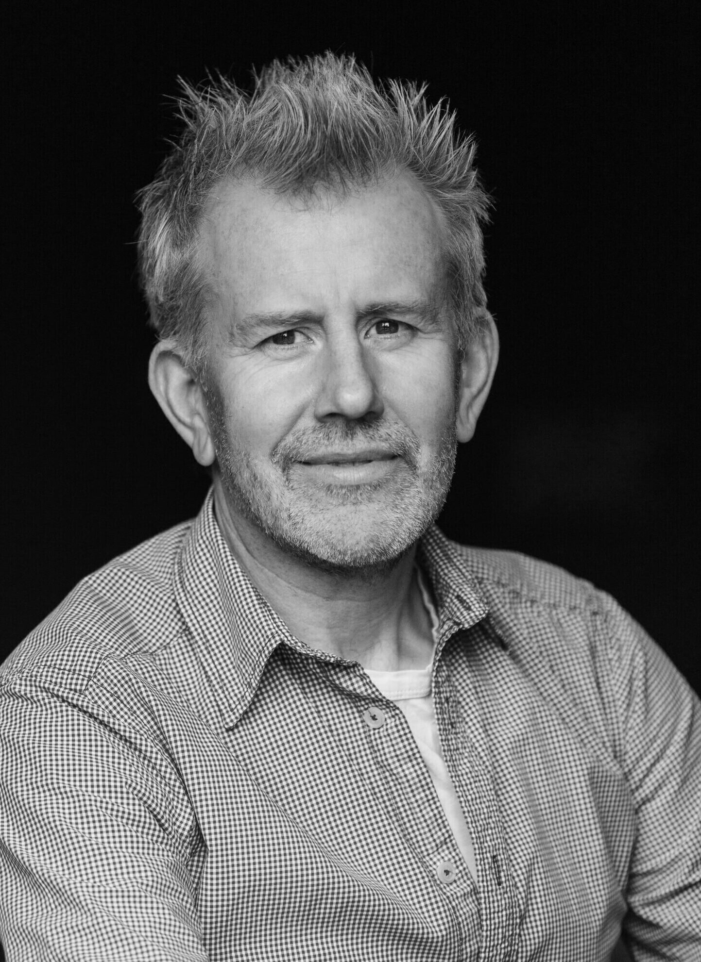 A man with spiky hair and a beard looks at the camera. He is wearing a collared shirt with a t-shirt underneath.