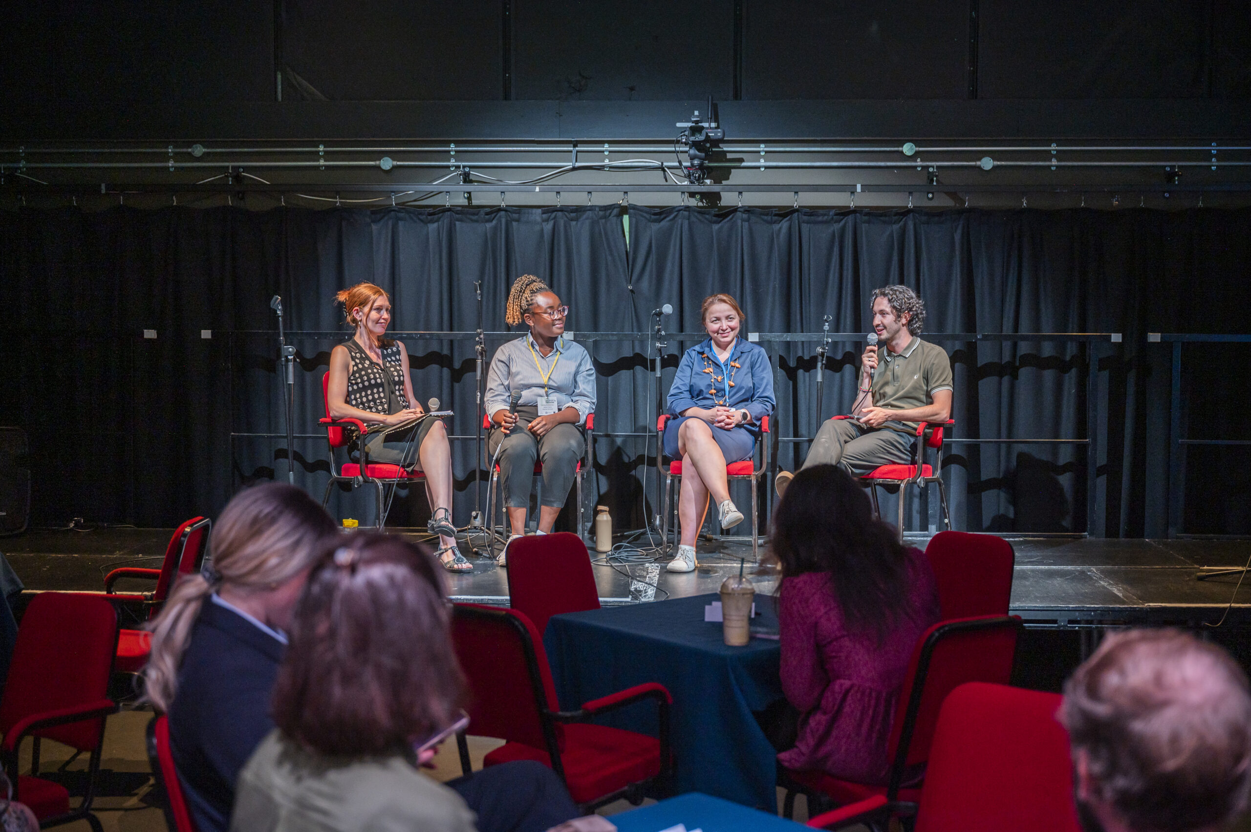 Four people sit in chairs on a stage. There are people sitting in chairs watching them. One of the people on the stage is holding a microphone and talking.