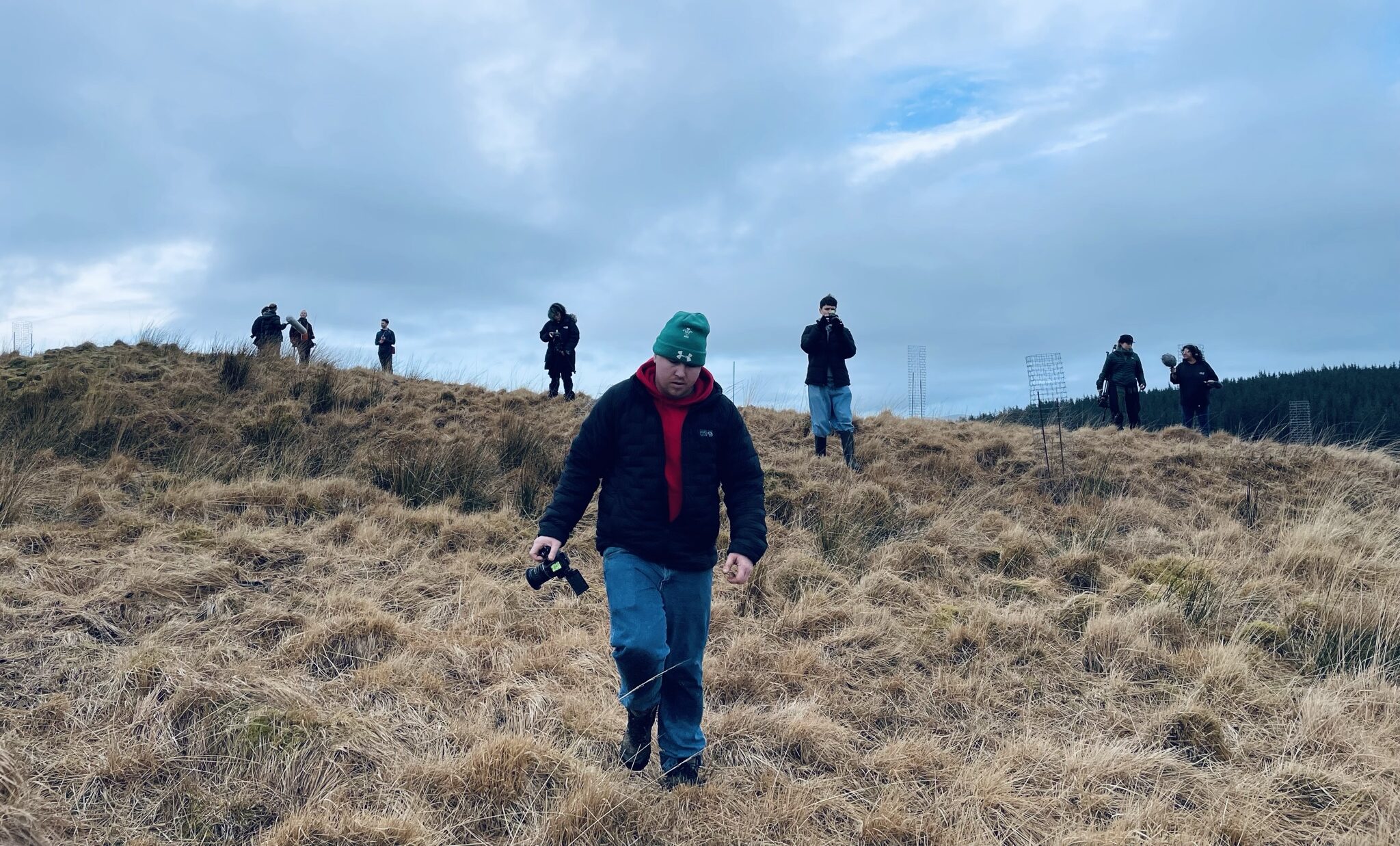 People in coats and hats walk down a hillside carrying filming equipment. The person in the foreground is carrying a camera and is taking a step forward.